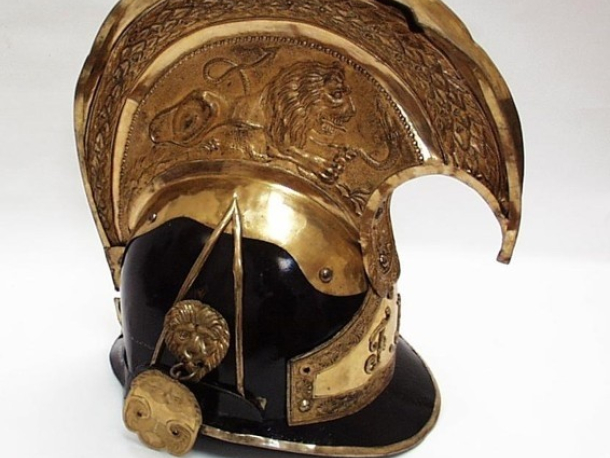 Rakouská důstojnická přilba jezdecká – zvon, z konce 19. století, na temeni vysoký hřeben z mosazného plechu, zdobený reliéfem souboje lva s hadem, po stranách masky lvů, vpředu znak s iniciálami Františka Josefa I.