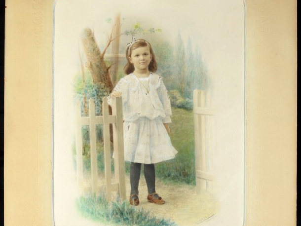 Děvče ve světlých šatech, tmavých punčochách a hnědých botách u bílého plůtku, fotografie kolorovaná v kartonové paspartě, foto: ing. J. Dědeček, v pravém dolním rohu signovaná: V. Jansa 1907 PRAHA