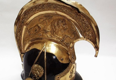 Rakouská důstojnická přilba jezdecká – zvon, z konce 19. století, na temeni vysoký hřeben z mosazného plechu, zdobený reliéfem souboje lva s hadem, po stranách masky lvů, vpředu znak s iniciálami Františka Josefa I.